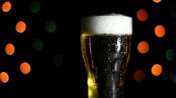 A glass with a cold light foamy beer stands on a table on a black background with colored lights. Drops of water flow down the glass. Mood of the holiday. HD video