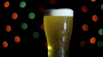 A glass of cold light foamy beer stands on a table on a black background with colored lights. Beer is foaming in a glass. HD video