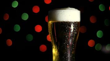 A glass with a cold light foamy beer stands on a table on a black background with colored lights. Drops of water flow down the glass. HD video