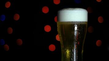 A glass of cold light foamy beer stands on a bar counter on a black background with colored lights. Beer is foaming in a glass. video