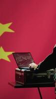 Vertical Chinese army military dispatcher looking at surveillance transmission on laptop, relaying informations for troops using headset, isolated over fluttering China flag background, camera A video