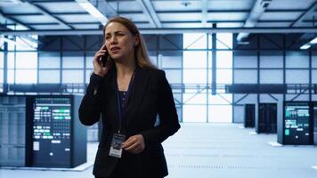Software engineer talking on smartphone in data center, coordinating server maintenance tasks. Woman surrounded by server racks and high tech equipment, speaking during telephone call, camera A video