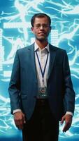 Vertical Smiling technician in startup stands with arms crossed, exuding professionalism and expertise. Portrait of cheerful Indian IT expert working in futuristic artificial intelligence workspace, camera A video