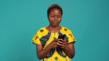 Native girl in ethnic outfit browsing internet webpages in studio, using her smartphone to check texts and social media platforms. Young person standing against blue background. Camera A. video