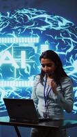Vertical Technician in high tech workspace managing call center tasks, speaking into headset while doing monitoring with laptop. AI company employee talking with clients through headphones mic, camera B video