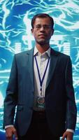 Vertical Upbeat engineer in startup doing advancements in AI field. Portrait of happy man standing with arms crossed in front of cutting edge display featuring AI and neural network graphics, camera B video