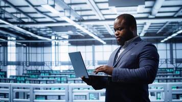 Programmer in data center typing on laptop, running error checking utilities. IT professional in server room looking for network issues affecting rigs performance using digital device, camera B video