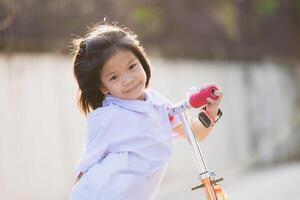 Portrait of smiling child plays with red scootering outdoors in a park, sunny day. Girl playing on suburban street on summer or spring time. Healthy outdoor activity. Cute kid on his way to school. photo