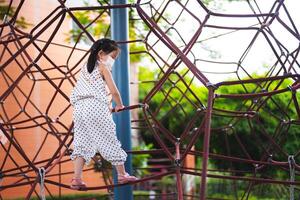 Girl having fun climbing a rope play equipment in playground. Child wear masks to protect her health. The play equipment is a rope that is woven into a three-dimensional shape for children to climb. photo