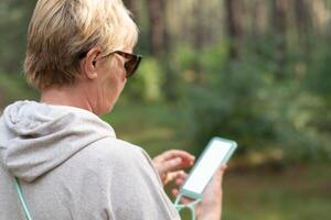 middle-aged woman in the forest takes daily steps, looks at the route map on her smartphone, turns on the pedometer, white screen mockup soft focus photo
