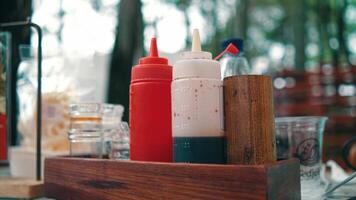 Condiment bottles with ketchup, mayonnaise, and soy sauce in a wooden holder on a restaurant table, with a blurred background video