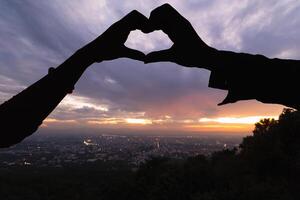 Silhouette of a pair of raised hands forming a heart shape on a beautiful evening landscape background Hand made heart symbol idea showing love friendship and kindness to couples and friends. photo