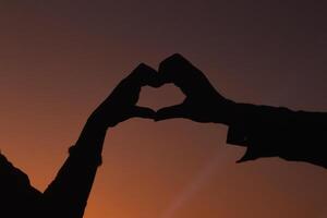 Silhouette of a pair of raised hands forming a heart shape on a beautiful evening landscape background Hand made heart symbol idea showing love friendship and kindness to couples and friends. photo