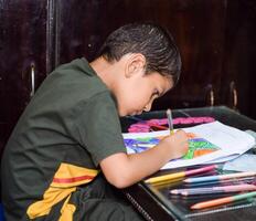 Smart Indian little boy drawing with colourful pencils kit of various colours during the summer vacations, Cute Indian Kid Sketching colourful drawing on wooden table photo