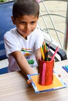Smart Indian little boy performing best out of waste activity with different colourful objects during the summer vacations, Cute Indian Kid doing best our of waste activity at home on wooden table photo