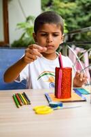 Smart Indian little boy performing best out of waste activity with different colourful objects during the summer vacations, Cute Indian Kid doing best our of waste activity at home on wooden table photo