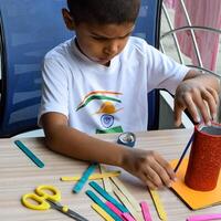 Smart Indian little boy performing best out of waste activity with different colourful objects during the summer vacations, Cute Indian Kid doing best our of waste activity at home on wooden table photo