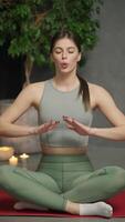 Vertical portrait of a calm brunette girl in a gray top and green sweatpants who sits in the lotus position on the red sports carpet and does yoga and breathing practices in a modern apartment video