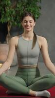 Vertical portrait of a happy brunette girl in a sports uniform sitting on a red mat and in a lotus position and doing yoga and sports in a modern apartment near lit candles and a houseplant video