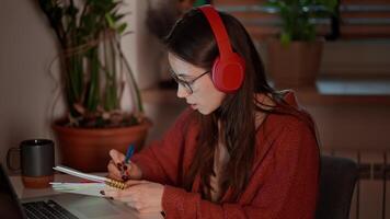 Confident brunette girl in wireless headphones and glasses takes notes in her notes while learning foreign languages using online lessons on a laptop while sitting in a modern kitchen in an apartment video