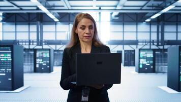 Software developer walking in data center, overseeing server operations using laptop. Woman ensuring optimal performance and data integrity in server farm using notebook, camera B video