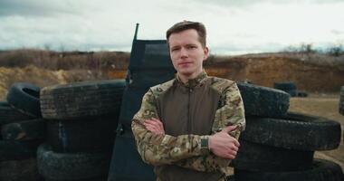 Portrait of a confident military man in a camouflage green uniform who folds his arms on his chest and poses with a serious expression near barricades made of car tires in the steppe during combat video
