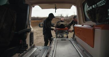View from the side of the ambulance cabin three military men put a wounded man in camouflage uniform on a snare in an ambulance while providing medical care on the battlefields video
