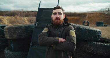 Portrait of a confident brunette man with a beard in a dark green army uniform who folds his arms on his chest and confidently poses near barricades made of car tires during a combat training exercise video