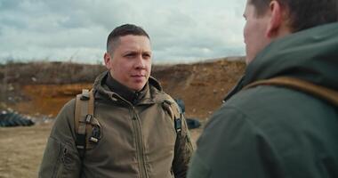 Over his shoulder a confident military man communicates with his colleague in a dark green army uniform and a backpack on his shoulders on the territory of a military training ground video