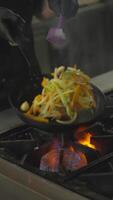 Close-up vertical of a confident chef in black gloves frying vegetables on a gas burner in the kitchen of a restaurant. And the cook fries vegetables in a frying pan turning them over and video