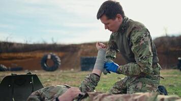 Side view of a confident male military man in a green camouflage uniform with medical gloves bandaging the hand with fabric bandages of a wounded soldier during a training exercise at a military video