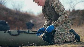 View from the side of a confident young male army medic in a camouflage green uniform strapping and stabilizing an unconscious man soldier on a medical army stretcher at an army training ground video