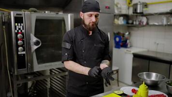 Confident male chef with a beard in a black uniform puts on gloves and starts preparing a salad in a modern kitchen in a restaurant. Start of the working day in the kitchen and use of protective video