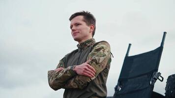 From the side a confident man with a short haircut in an army camouflage uniform stands confidently against the sky and looks to the side with his arms folded on his chest outside the city video