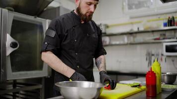 Close up of a confident male chef in a black uniform with a bandana and protective gloves cutting a salad and putting it into a bowl while preparing a dish in a modern kitchen in a restaurant video