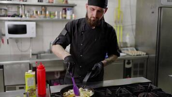 Confident male chef with a beard in a black uniform and with black protective gloves fries vegetables in a frying pan adding oil in a modern kitchen in a restaurant video