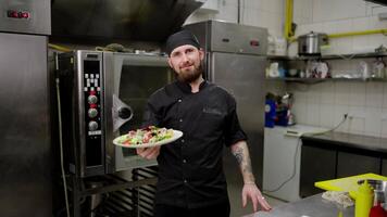 Portrait of a confident Happy male chef in a black uniform and with a beard who holds his ready-made salad in his hands before serving in a restaurant in the kitchen video