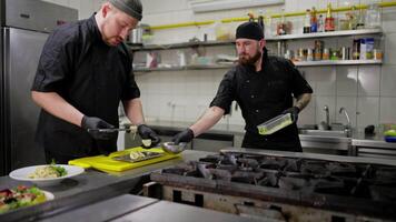 A male trainee chef in a black uniform prepares a dish with the help of an experienced chef in the kitchen in a restaurant. A male experienced chef helps a trainee to prepare a dish correctly and use video