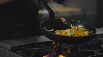 Close-up a professional chef in a black uniform mixes thinly sliced vegetables on a hot frying pan while preparing a dish on a gas burner in a restaurant kitchen video