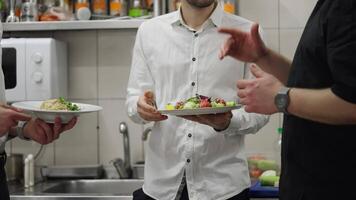 Close-up of a cook and two waiters communicating about dishes while working in the kitchen in a restaurant. Two waiters in shirts hold salads in their hands and communicate with the chef about their video