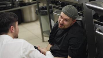 A confident professional chef in a black uniform with a beard communicates with a young guy waiter who supports and pats the chef on the shoulder while sitting on the floor in the kitchen in a video