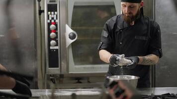 Confident professional chef in a black uniform with a beard places dough on the table with flour and rolls it out with his hands while preparing pizza in the kitchen in a restaurant video
