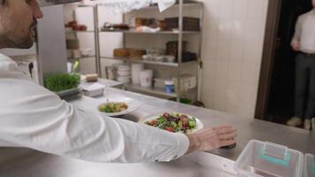 A male cook in a white shirt puts ready-made dishes and salads on the table, which he notifies using a bell and the waiter takes the dish taking them into the restaurant hall video