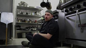 Portrait of a very tired man with a beard of a professional chef in a black uniform who sits on the floor leaning on the table in the kitchen of a restaurant and sighs heavily video