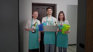 Portrait of a happy trio of cleaners in blue fronts with various tools and a cleaner for cleaning in their hands two men and a brunette girl in a modern apartment on a cleaning call video