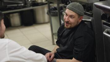 Close up over the shoulder of a confident and happy cook with a beard in a black uniform talking to a waiter in a white shirt while sitting on the floor in the kitchen in a restaurant video