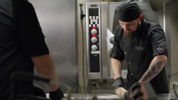 Professional chef in black uniform and bandana rolls out pizza dough using a wooden rolling pin in the kitchen in a restaurant. Cooperation of chefs in preparing dishes in the kitchen video