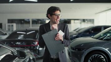 A happy and cheerful confident brunette man in glasses and a business suit an assistant in a car dealership with a tablet in his hands walks along the cars and dances video