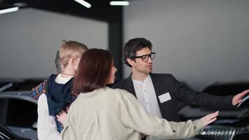 Confident brunette assistant guy in a business suit communicates with a family of clients in a modern car showroom. Over the shoulder a confident brunette girl along with her husband and little son video