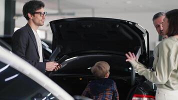 A confident man in glasses and an suit with a tablet in his hands communicates with a brunette girl with her husband and little son about a specific car model near an open trunk in a car showroom video